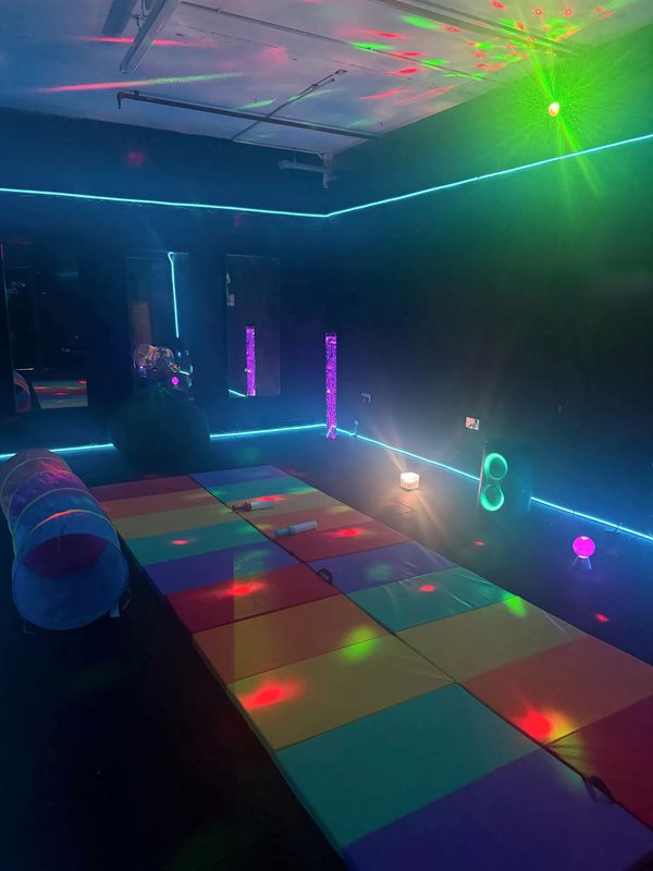 Sensory room with green and red lights dancing across the ceiling, a rainbow of soft floor mats and a glowing bubble tube